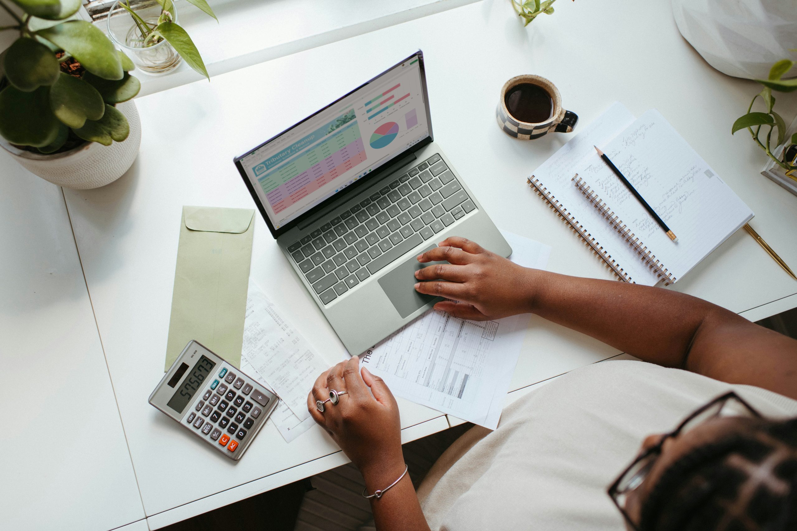 Person working at a desk with a laptop, calculator and notebook while reviewing financial information.