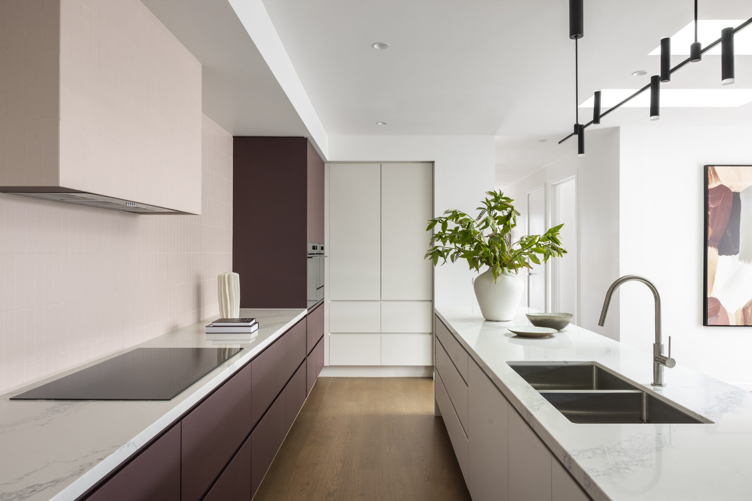 Contemporary kitchen featuring contrasting burgundy and light-toned cabinets, marble-look benchtops, a double-basin sink, induction cooktop, and a large potted plant on the island. Skylights and black linear pendant lights brighten the space.