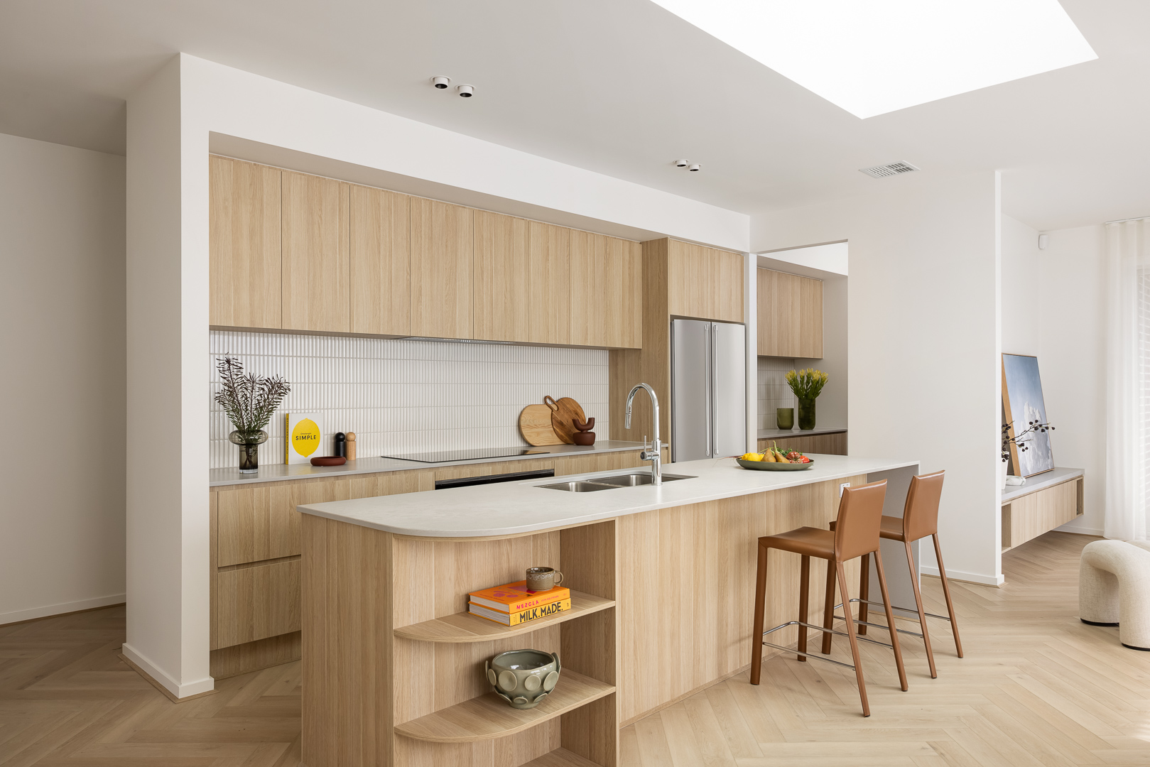 Contemporary kitchen featuring light oak cabinets, a white stone island with built-in shelving and sink, stainless steel refrigerator, two tan bar stools, and decorative items. The space has herringbone wood flooring, white walls, and abundant natural light from a skylight.