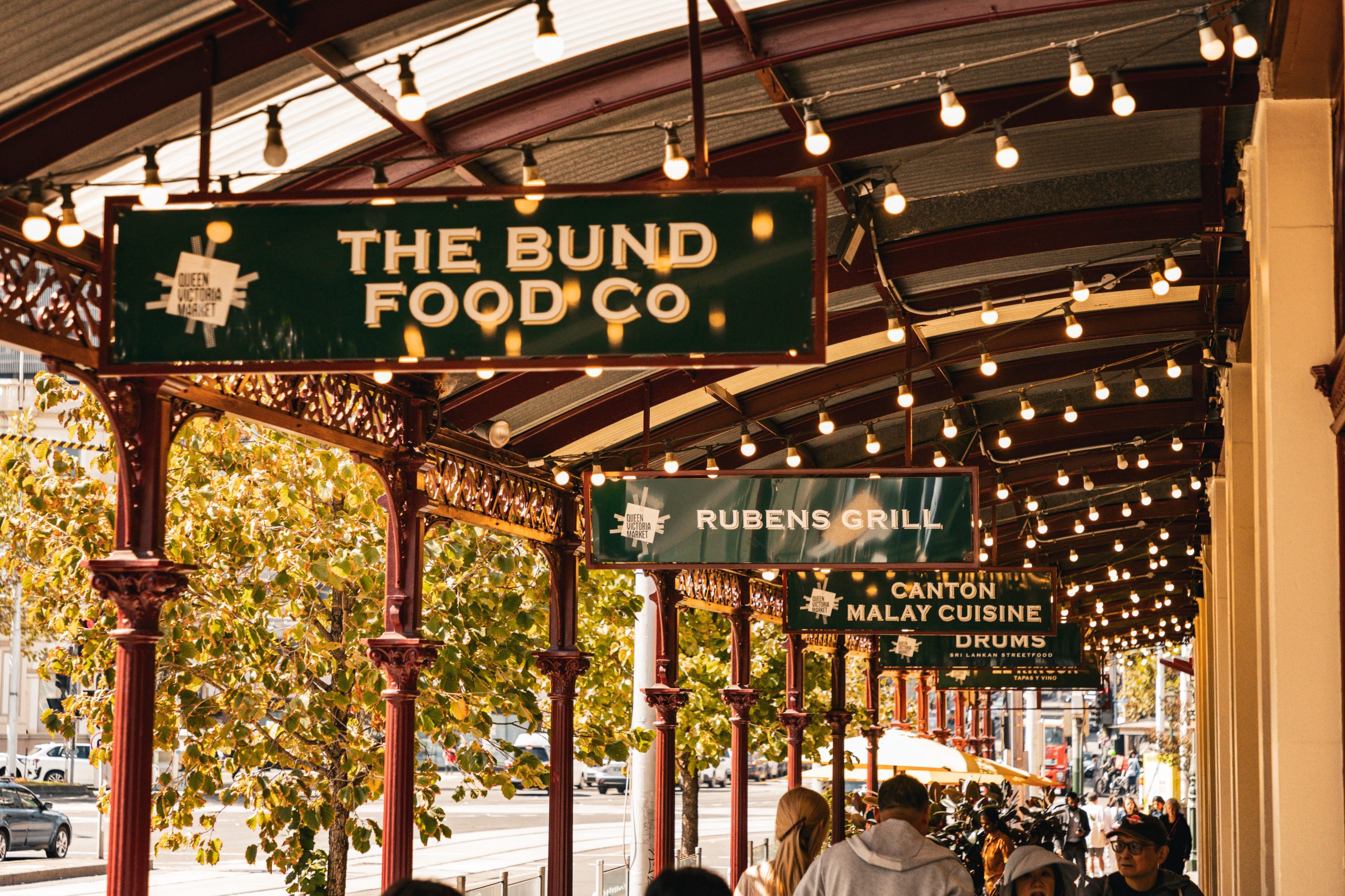 Covered market walkway lined with decorative iron columns and warm string lights, featuring hanging restaurant signs for The Bund Food Co, Rubens Grill, Canton Malay Cuisine and Drums, with trees and pedestrians visible along the street.